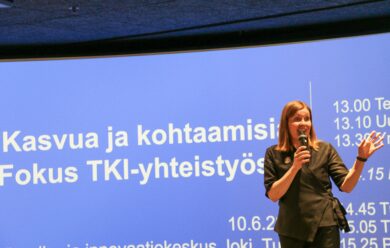 Nainen puhumassa mikrofonin kanssa lavalla TKI-yhteistyötä käsittelevässä seminaarissa. Taustalla sininen esitysdia, jossa lukee 'Kasvua ja kohtaamisia – Fokus TKI-yhteistyössä' sekä tapahtuman aikataulu ja paikka, Turun tiede- ja innovaatiokeskus Joki 10.6.2024.