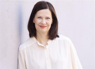 Minna Arve seisoo vaaleaa seinää vasten, katsoo kameraan ja hymyilee. Hänellä on päällään vaalea pusero ja tumma hame, rennon ja asiantuntevan oloinen ilme.