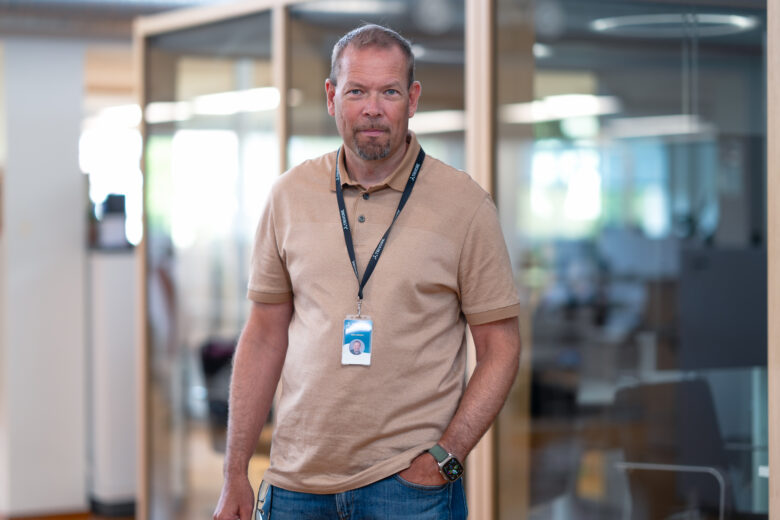 CEO Mika Aromaa standing in a modern office environment, looking directly at the camera. He is wearing a light brown polo shirt and a black TRIVORE lanyard with an ID badge. In the background, there are glass-walled meeting rooms and softly lit office interior.