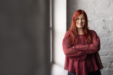 Unna Ahokas seisoo hymyillen ikkunan vieressä kädet ristissä. Hänellä on pitkät punaiset hiukset ja viininpunainen pusero. Taustalla vaalea tiiliseinä ja ikkunasta tuleva valo luo pehmeän tunnelman.