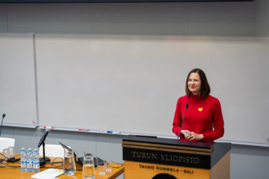 Business Turun toimitusjohtaja Minna Arve puhumassa Turun yliopiston Tauno Nurmela -salissa. Hän seisoo puhujakorokkeen takana punaisessa puserossa mikrofonin edessä.